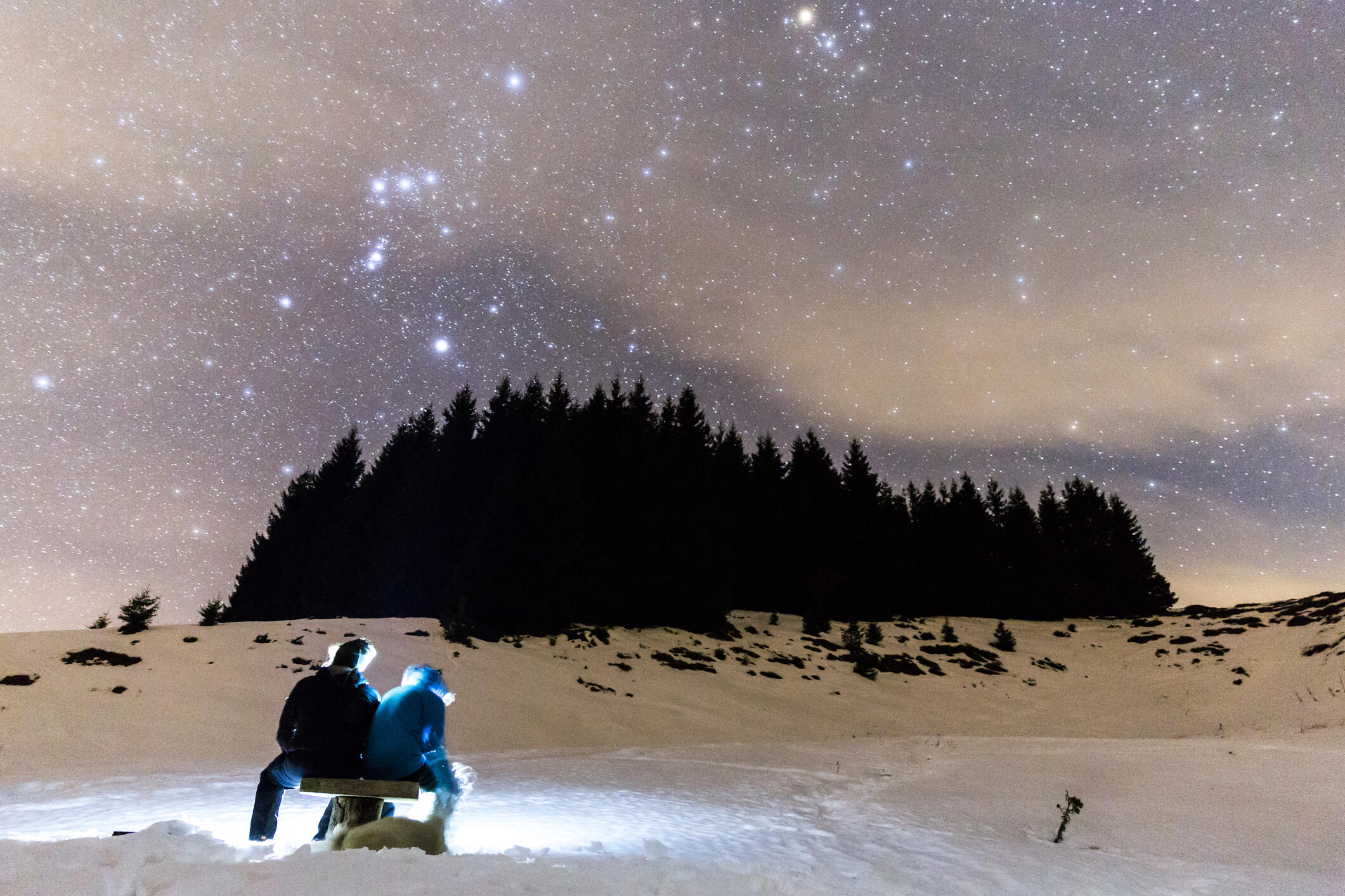 The Milky Way over the winter mountain landscape with pine trees. Two people sitting in the foreground watching the falling stars.