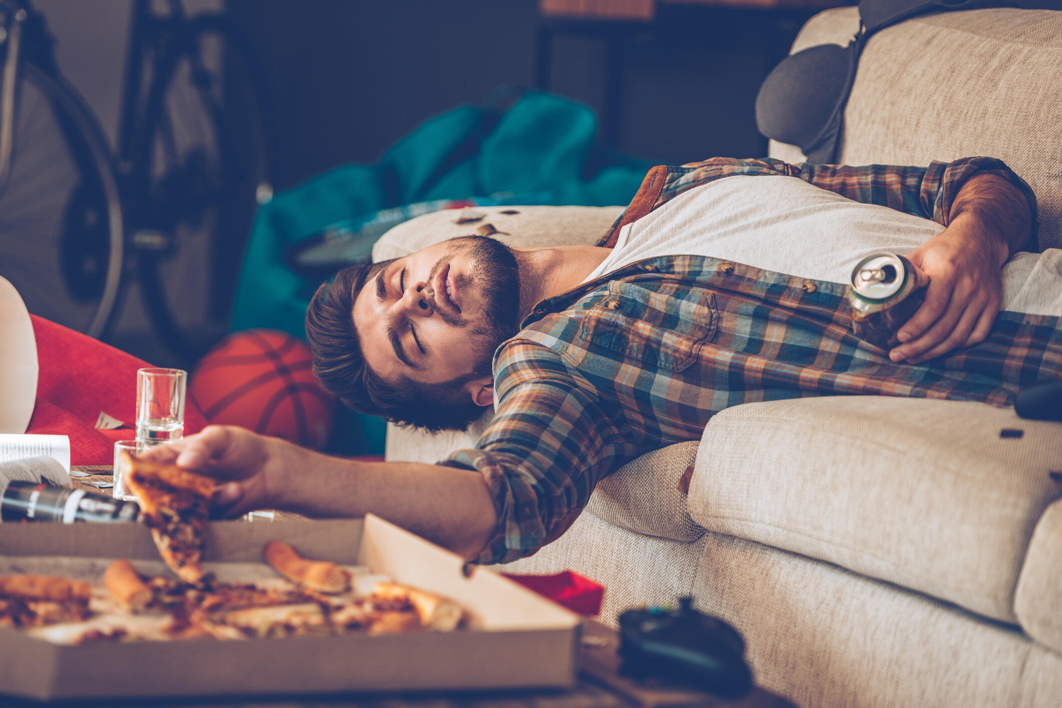 Young handsome man passed out on sofa with pizza slice and beer can in his hand in messy room after party