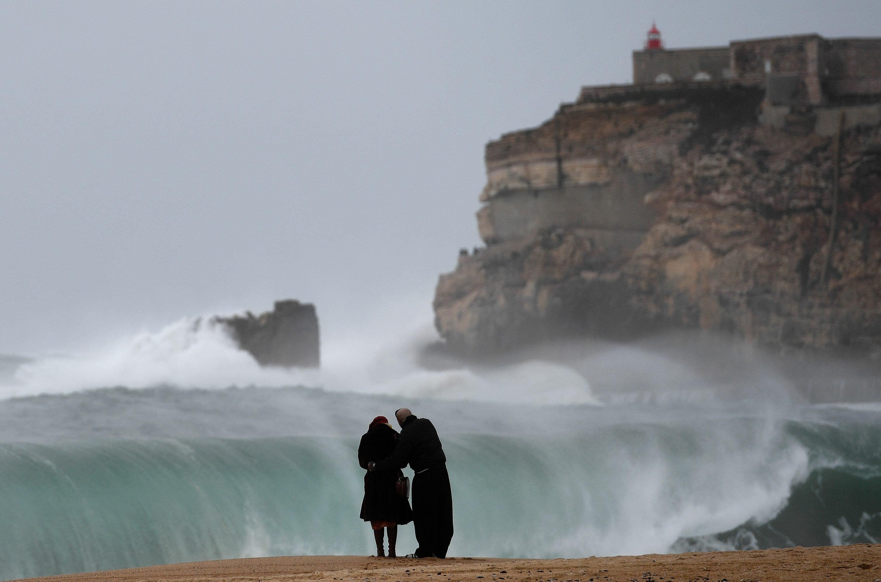 TOPSHOT - A couple stands on the beach as big waves break near the Sao Miguel Arcanjo fort in Nazare, central Portugal, on February 2, 2017. / AFP PHOTO / FRANCISCO LEONG        (Photo credit should read FRANCISCO LEONG/AFP/Getty Images)