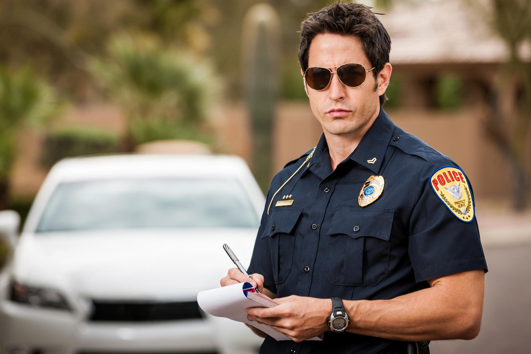 A mid-adult caucasian male police officer stands in front of a car, writing a ticket for a moving violation.