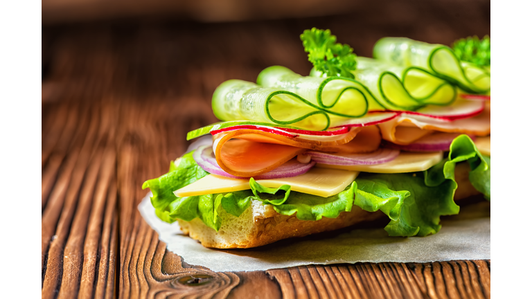 close up of sandwich with ham, cheese, bacon, radish, lettuce, cucumbers and onions on paper, wooden background