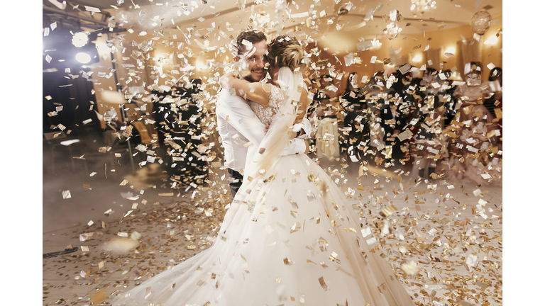 Gorgeous bride and stylish groom dancing under golden confetti at wedding reception. Happy wedding couple performing first dance in restaurant. Romantic moments