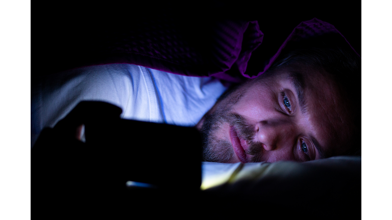 Bearded young man is lying in bed under his blanket looking at phone