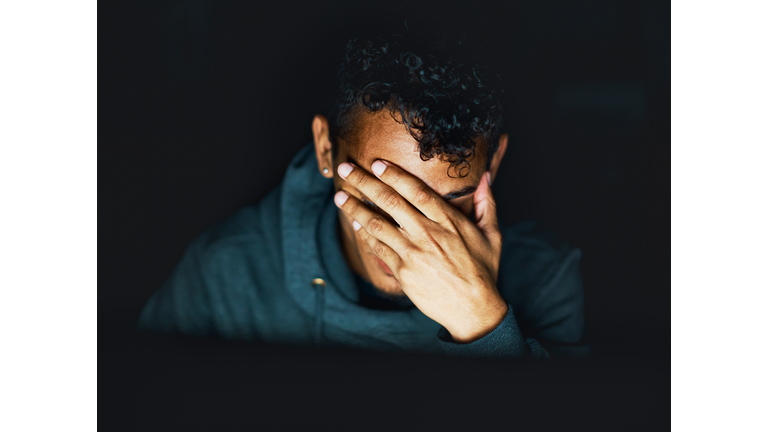 Young man sitting in the dark covers his face