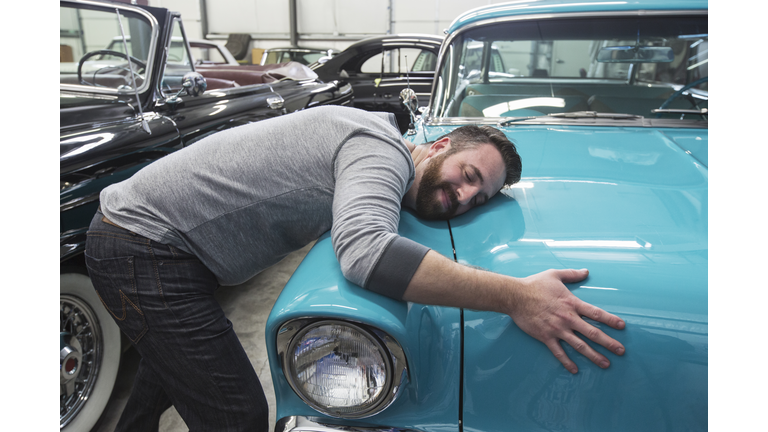 A Caucasian male hugging the hood of his old sedan in a classic car repair shop.