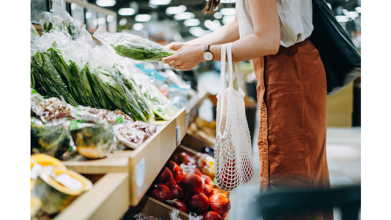 Cropped shot of young Asian woman shopping for fresh organic groceries in supermarket. She is shopping with a cotton mesh eco bag and carries a variety of fruits and vegetables. Zero waste concept