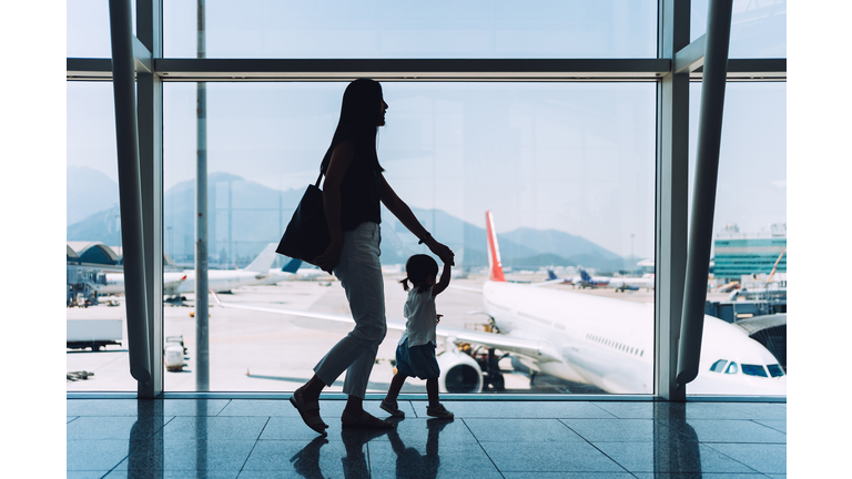 Silhouette of joyful young Asian mother holding hands of cute little daughter looking at airplane through window at the airport while waiting for departure