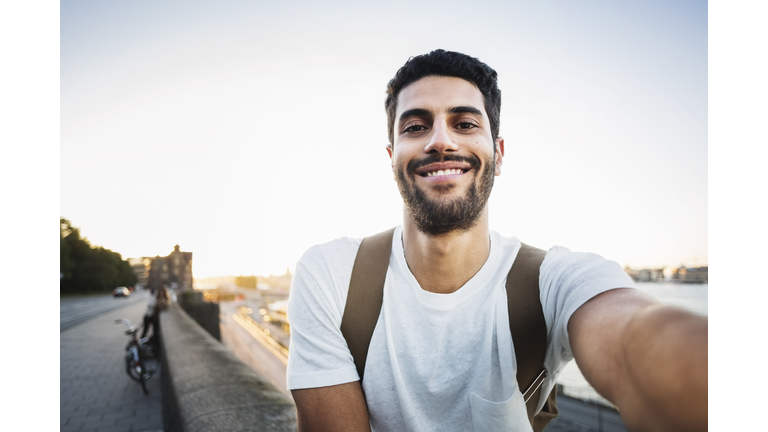 Portrait of happy male tourist sitting on retaining wall of bridge