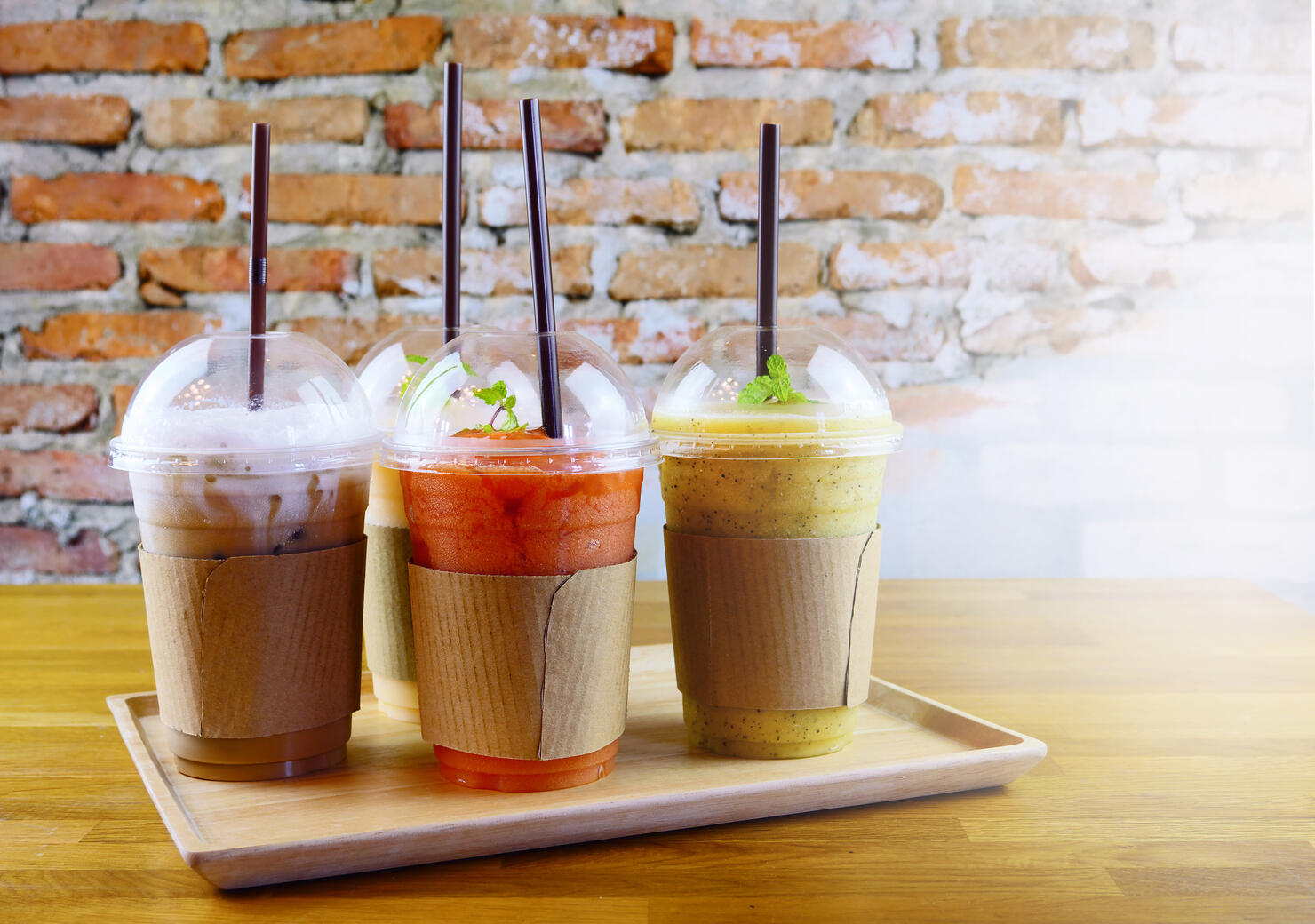 Smoothies and iced coffee in plastic cup on wooden tray and brick wall background in cafe. Take away drinks concept.