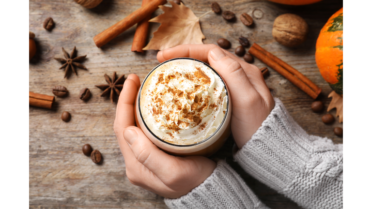 Woman holding glass of tasty pumpkin spice latte on wooden table, flat lay composition