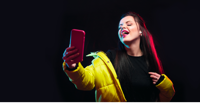 Tik tok advertisement concept.Young girl posing with smart phone in her hands, making selfie on black background.TIK TOK is a popular social network on the internet.Studio shots, copy space for text