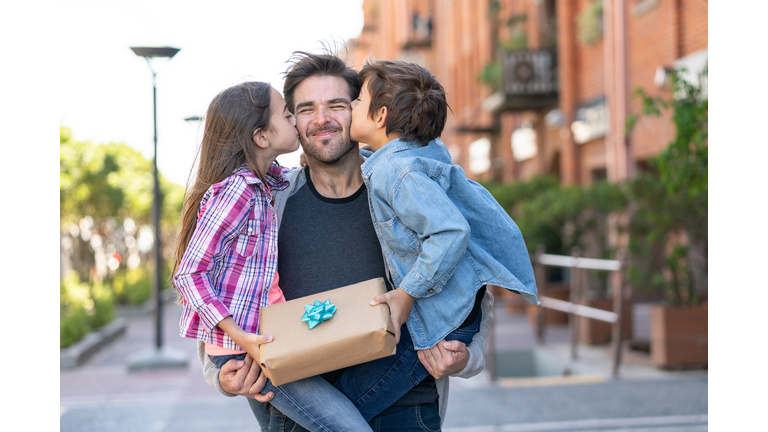 Loving son and daughter kissing daddy on the cheek while he carries them both with a present for father's day