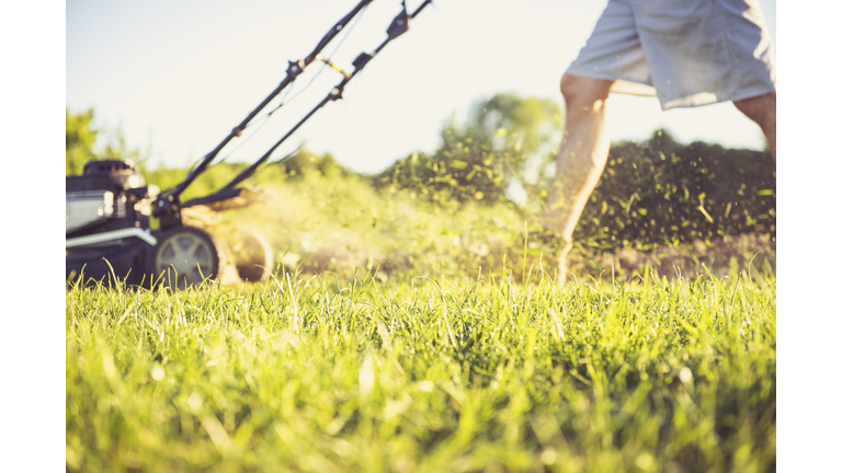 Low section of man mowing grassy field in yard against sky during sunset