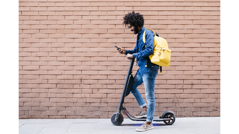 Young man with backpack and E-Scooter standing in front of brick wall looking at cell phone