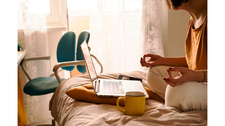 A middle-aged woman in white jeans and a yellow sweater sitting on the bed in a yoga pose in front of a laptop and a cup of coffee