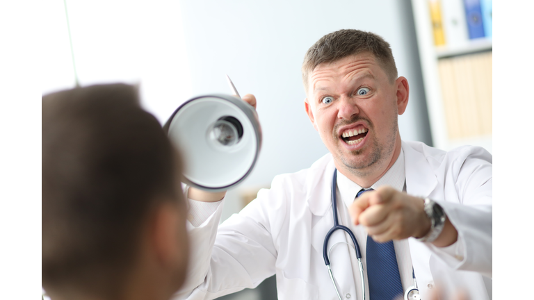 Angry doctor interrogates the patient, yelling at him, pointing to man he talking to and holding lamp base.