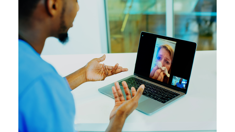 Portrait of a sick patient coughing into tissue being helped via tele medicine by a male doctor wearing blue scrubs uniform using laptop
