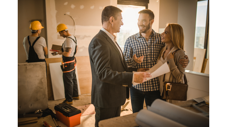 Happy couple and building contractor talking about housing plan at construction site.