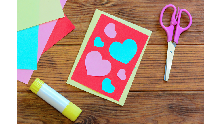 Valentines day or Mothers day postcard with pink and blue hearts. Scissors, glue stick, colored paper sheets set on a wooden background. Simple craft work with paper for children. Top view