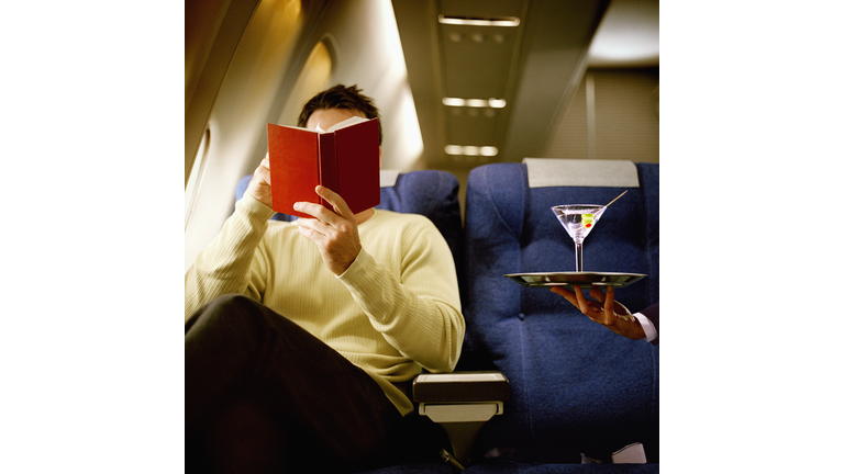 Man reading book, being served martini, in first class on airliner