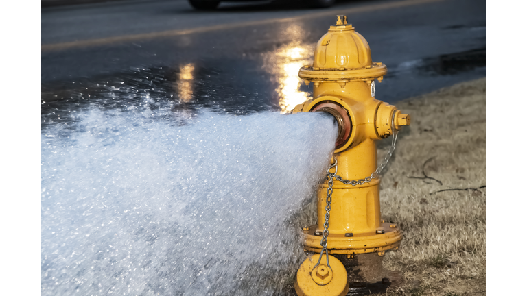 Close-up of yellow fire hydrant gushing water across a street with wet highway and tire from passing car behind