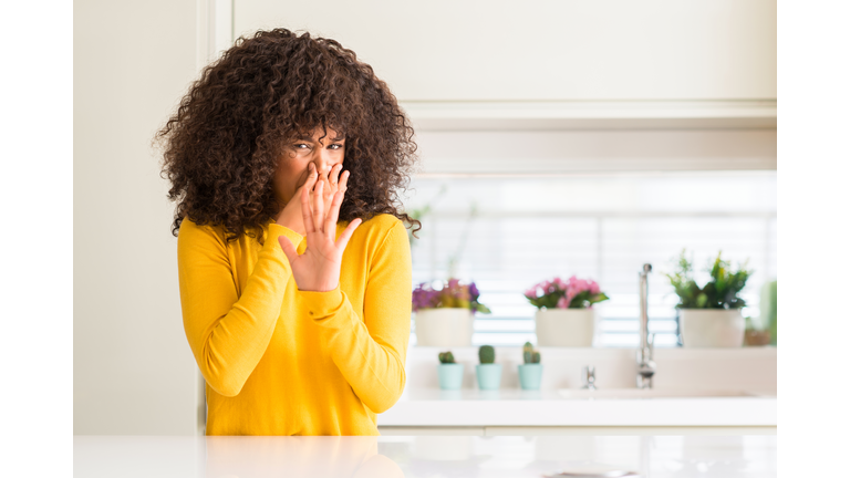 African american woman wearing yellow sweater at kitchen smelling something stinky and disgusting, intolerable smell, holding breath with fingers on nose. Bad smells concept.