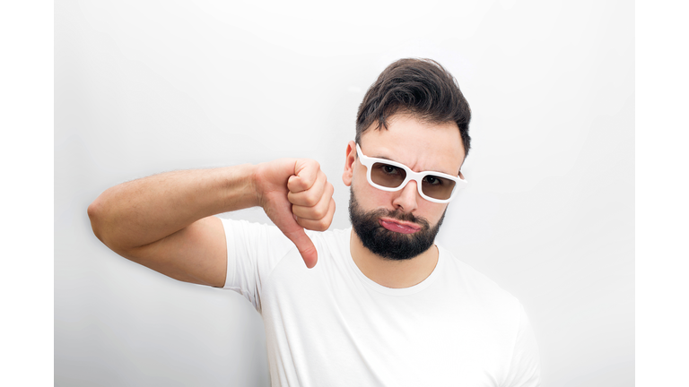 Sad and unhappy bearded young man stand and hold his big thumb down. He dislikes movie. Guy keeps cheeks blowed. Isolated on white background,