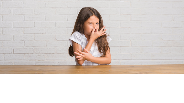 Young hispanic kid sitting on the table at home smelling something stinky and disgusting, intolerable smell, holding breath with fingers on nose. Bad smells concept.