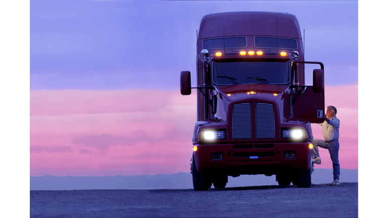 A silhouette of a truck driver getting into the cab of his commercial Class 8 truck tractor at sunrise.