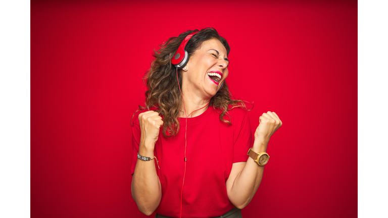 Middle age senior woman wearing headphones listening to music over red isolated background very happy and excited doing winner gesture with arms raised, smiling and screaming for success. Celebration concept.