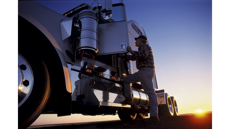 A silhouette of a truck driver getting into the cab of his commercial Class 8 truck tractor at sunrise.