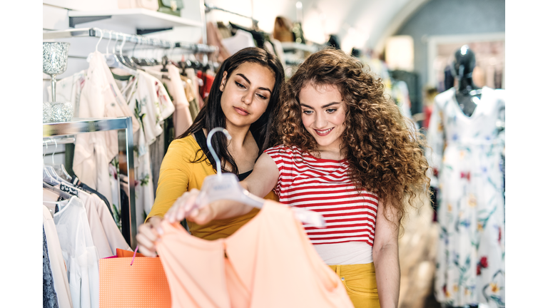 Two female teenager friends standing inside in the shop, holding and looking at dress.