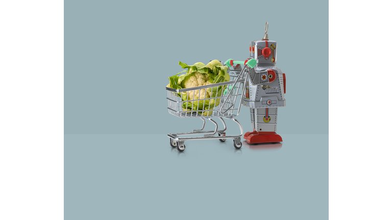 Toy robot pushing miniature shopping trolley with cauliflower against blue background