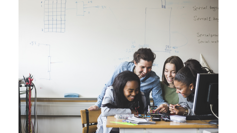 Smiling female teacher and high school teenage students preparing robot on desk in classroom