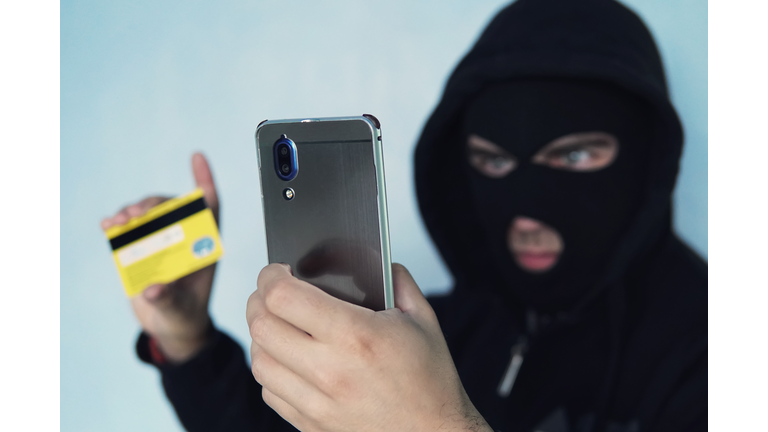 The thief scammer is holding a mobile phone and credit card. The concept of Bank card fraud through a virus in the smartphone. a thug in a black Balaclava and a black hood.