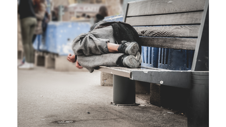 Poor homeless man or refugee sleeping on the wooden bench on the urban street in the city, social documentary concept, selective focus