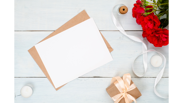 Flat lay blank card, craft paper envelope, red rose flowers and gift box. Top view wedding invitation card mockup. Send a postcard or mail letter concept.
