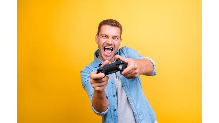 Portrait of young, cheerful, attractive, very excited guy holding joystick and playing video games over yellow background