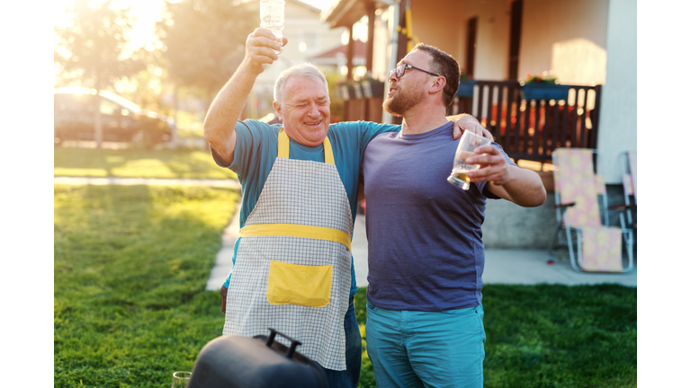 Cheerful father in law and son in law hugging and drinking beer while standing next to grill in backyard. Family gathering concept.