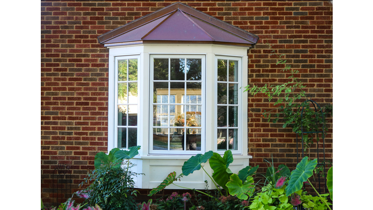 Bay window in a brick house with reflection of trees and view of windows and flowers inside and flowers and elephant ears outside