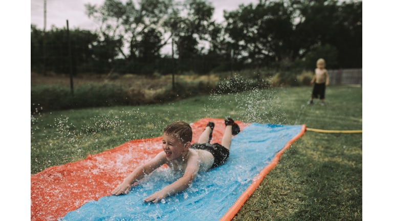 Boy sliding on water slide at yard with brother standing in background