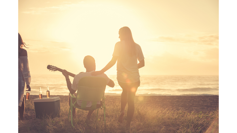 Young couple on beach looking at sunset with guitar.