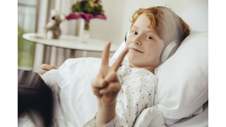 Sick boy lying in hospital making victory sign, wearing head phones