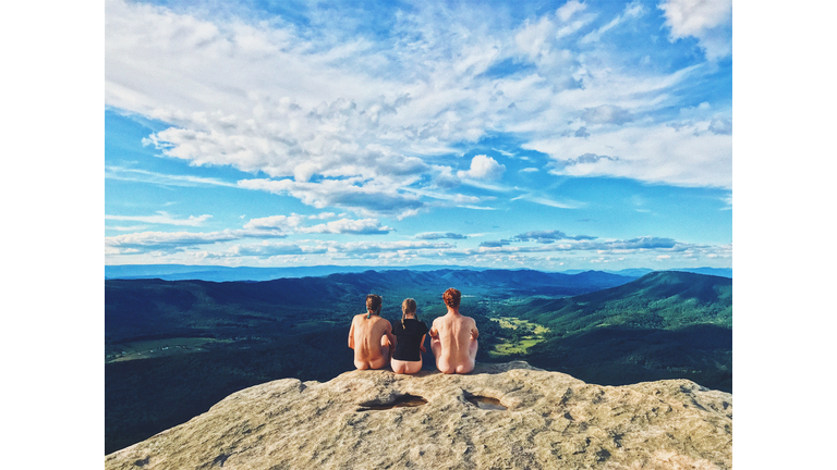 Rear View Of Semi Dressed Women With Naked Men Sitting On Cliff Against Sky