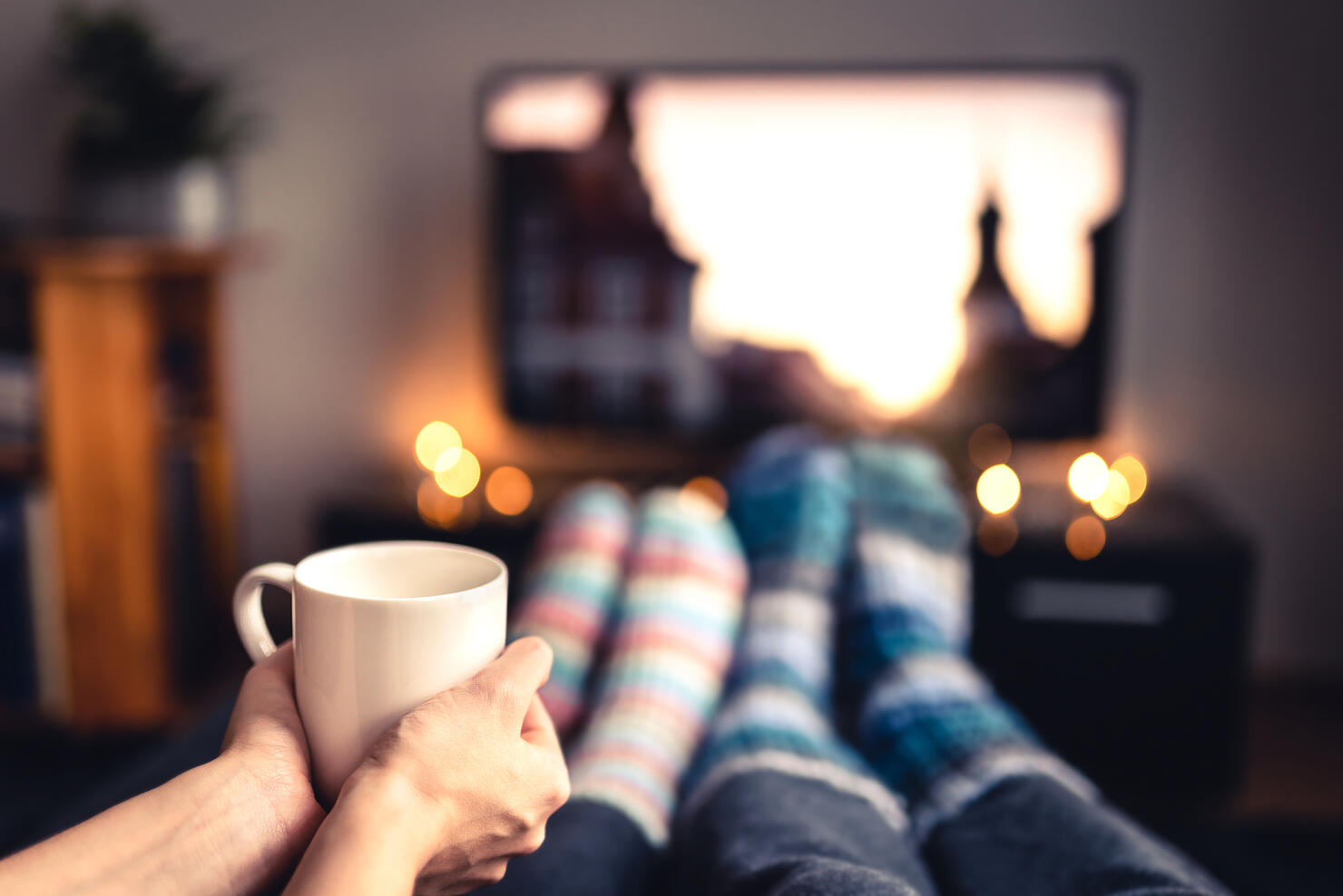 Couple drinking tea, hot chocolate, eggnog or mulled wine and watching tv in warm cozy woolen socks in winter. Woman holding cup of morning coffee in home living room. Sick people with flu.