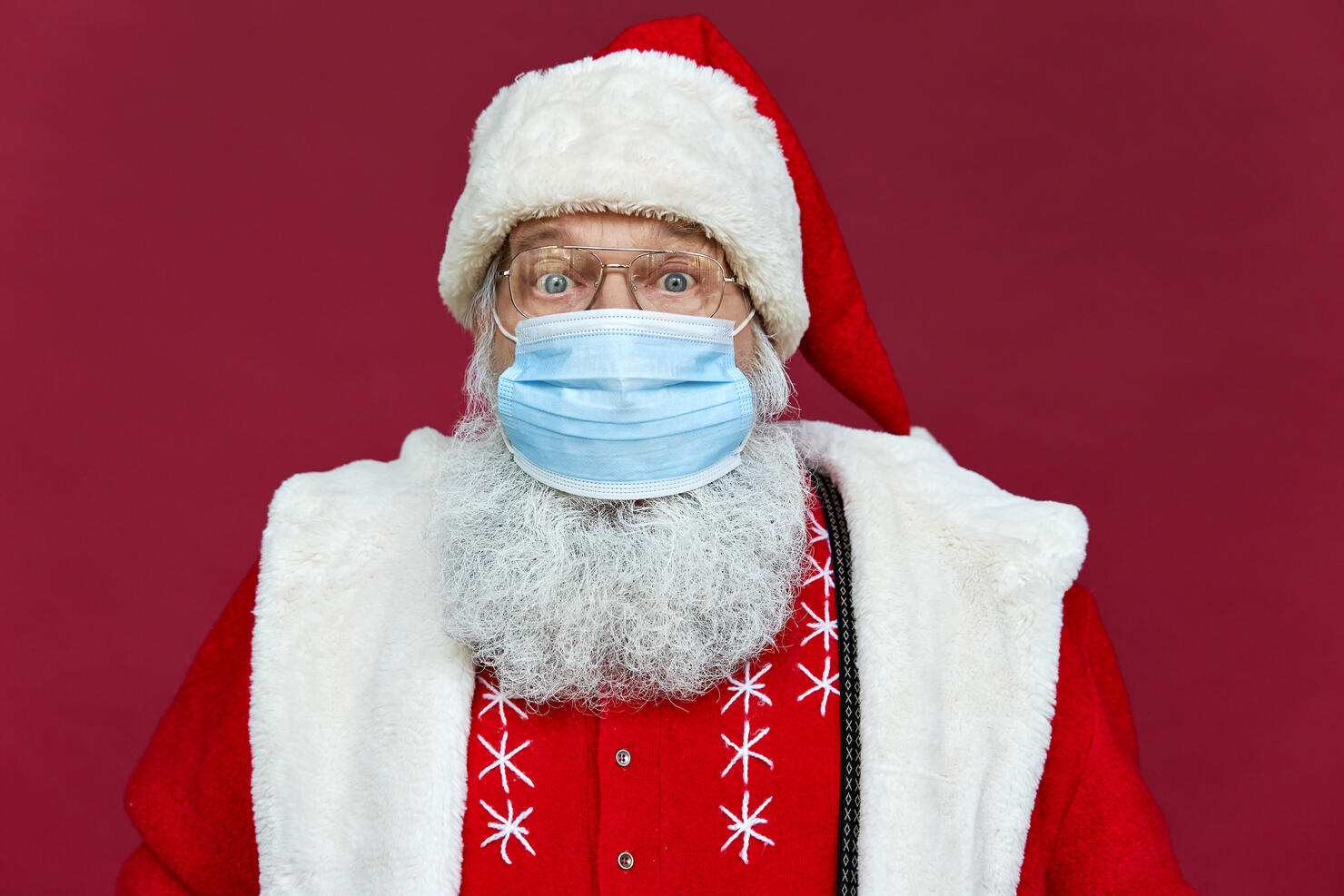 Close up portrait of funny old bearded surprised Santa Claus wearing costume, glasses, face mask looking at camera standing on Christmas red background. Covid 19 coronavirus safety protection concept.
