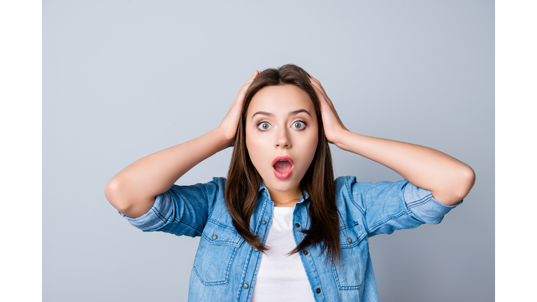 Omg! Close up portrait of amazed girl with wide open mouth and eyes in casual wear, holding head with her hands, wondering what happened, looking confused, lost on the grey background