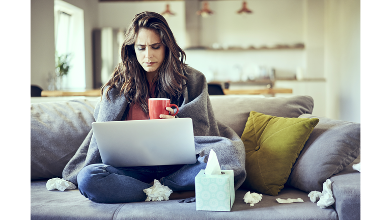 Sick woman sitting on sofa covered in blanket with cup of tea and laptop