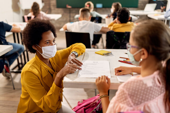 Black teacher with a face mask explaining exam results to elementary student in the classroom.