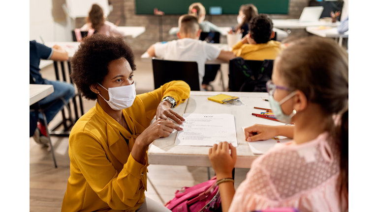 Black teacher with a face mask explaining exam results to elementary student in the classroom.